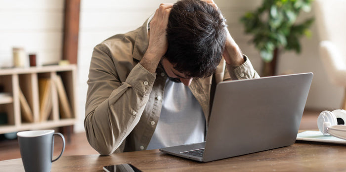 A tired man sitting at a desk with his laptop, leaning his head on his hand, appearing exhausted and stressed, with a cluttered workspace in the background.